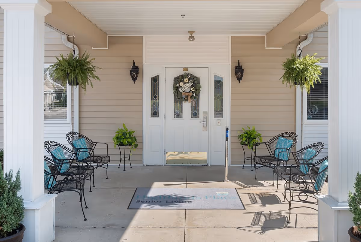Covered entrance area of Highlands Square senior living facility with white double doors decorated with a wreath, black metal chairs with blue cushions arranged on both sides, hanging green plants, and potted plants near white columns.