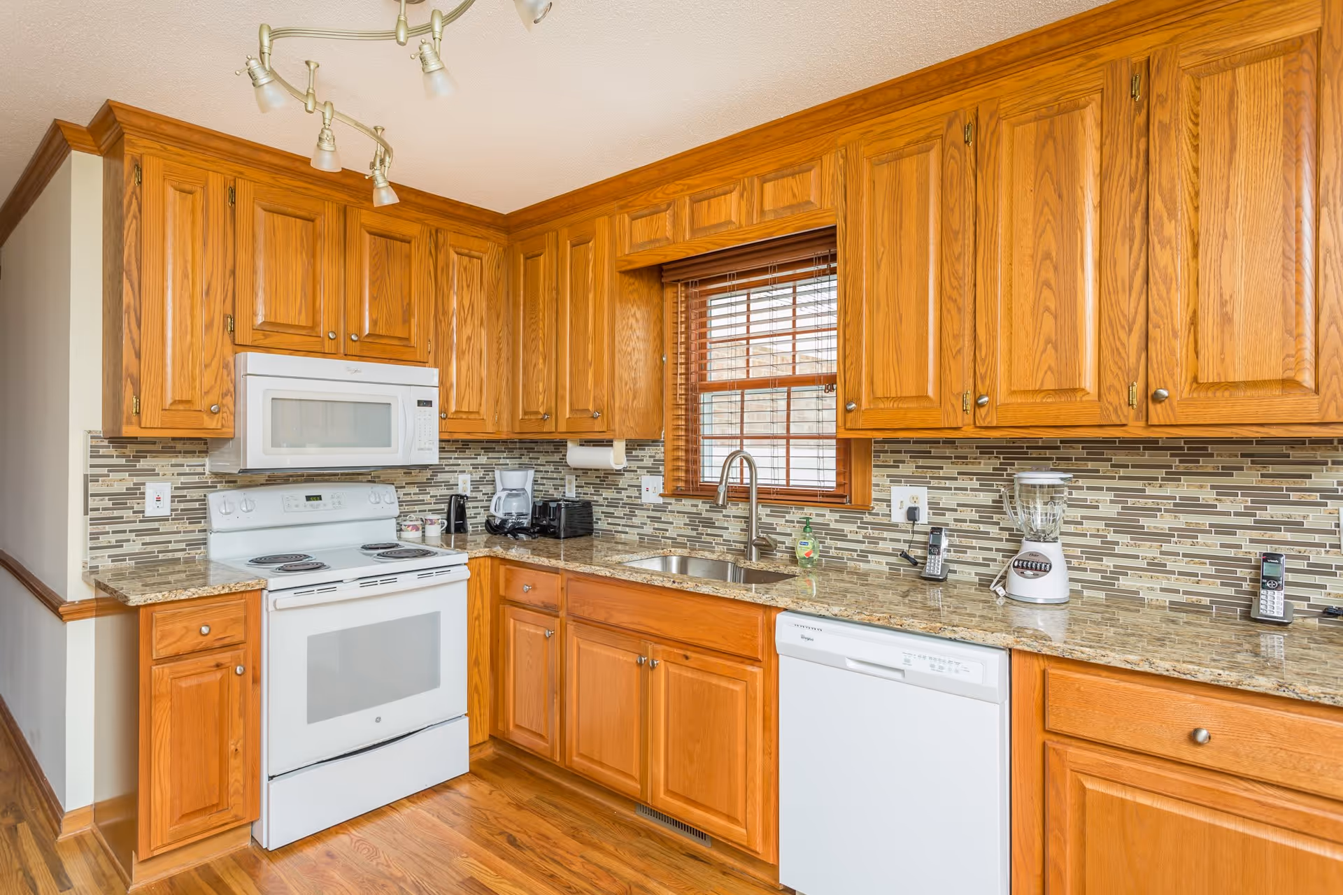 A bright kitchen with wooden cabinets, a white electric stove with an overhead microwave, a double sink under a window with wooden blinds, a dishwasher, and various small appliances on the granite countertop including a blender, coffee maker, and cordless phones. The backsplash features a mosaic tile design in neutral tones.