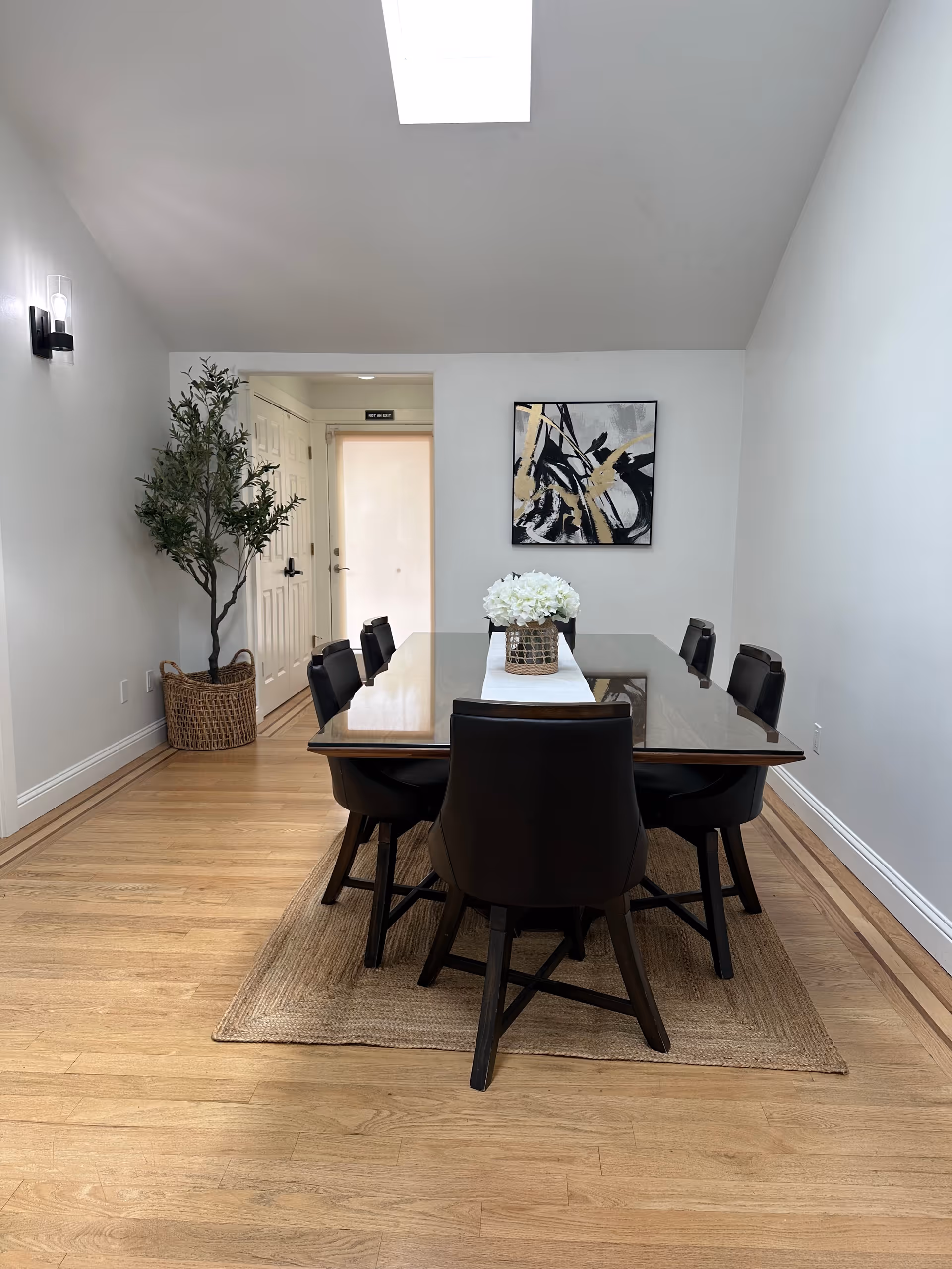 A dining room with a rectangular glass-top table surrounded by six black chairs. A white floral centerpiece sits on a white runner on the table. The room has light wood flooring with a woven rug under the table. There is a potted plant in a wicker basket to the left near a door, and an abstract black, white, and gold painting hangs on the wall. The ceiling has a skylight, and the walls are painted white.