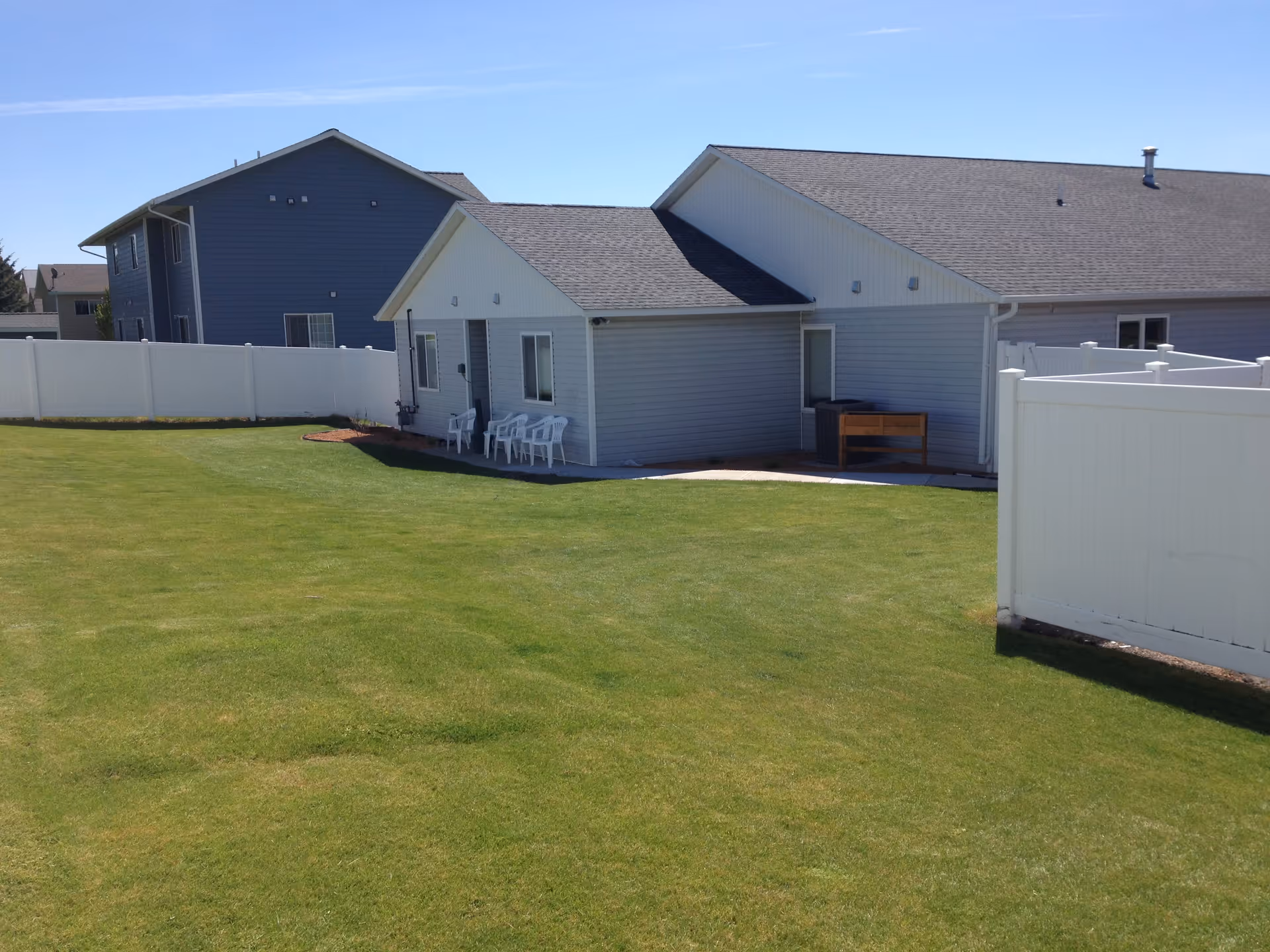 A single-story building with light gray siding and a dark gray roof, surrounded by a white fence. There are several white plastic chairs placed outside near the building. The area has a well-maintained green lawn under a clear blue sky.
