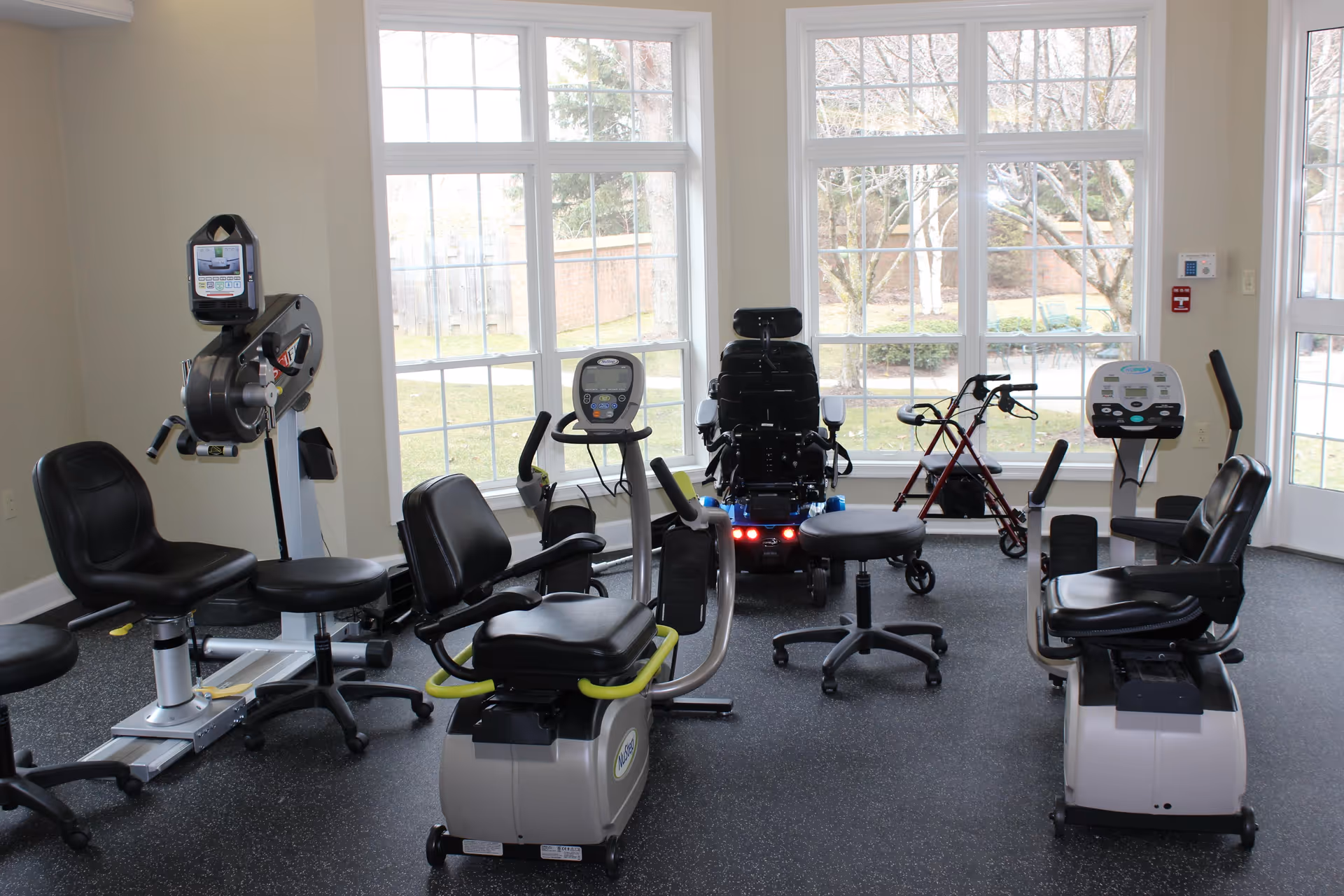 A bright exercise room with large windows showing an outdoor view. The room contains several pieces of exercise equipment including stationary bikes, a recumbent bike, an electric wheelchair, and a walker. The floor is covered with dark rubber gym flooring.