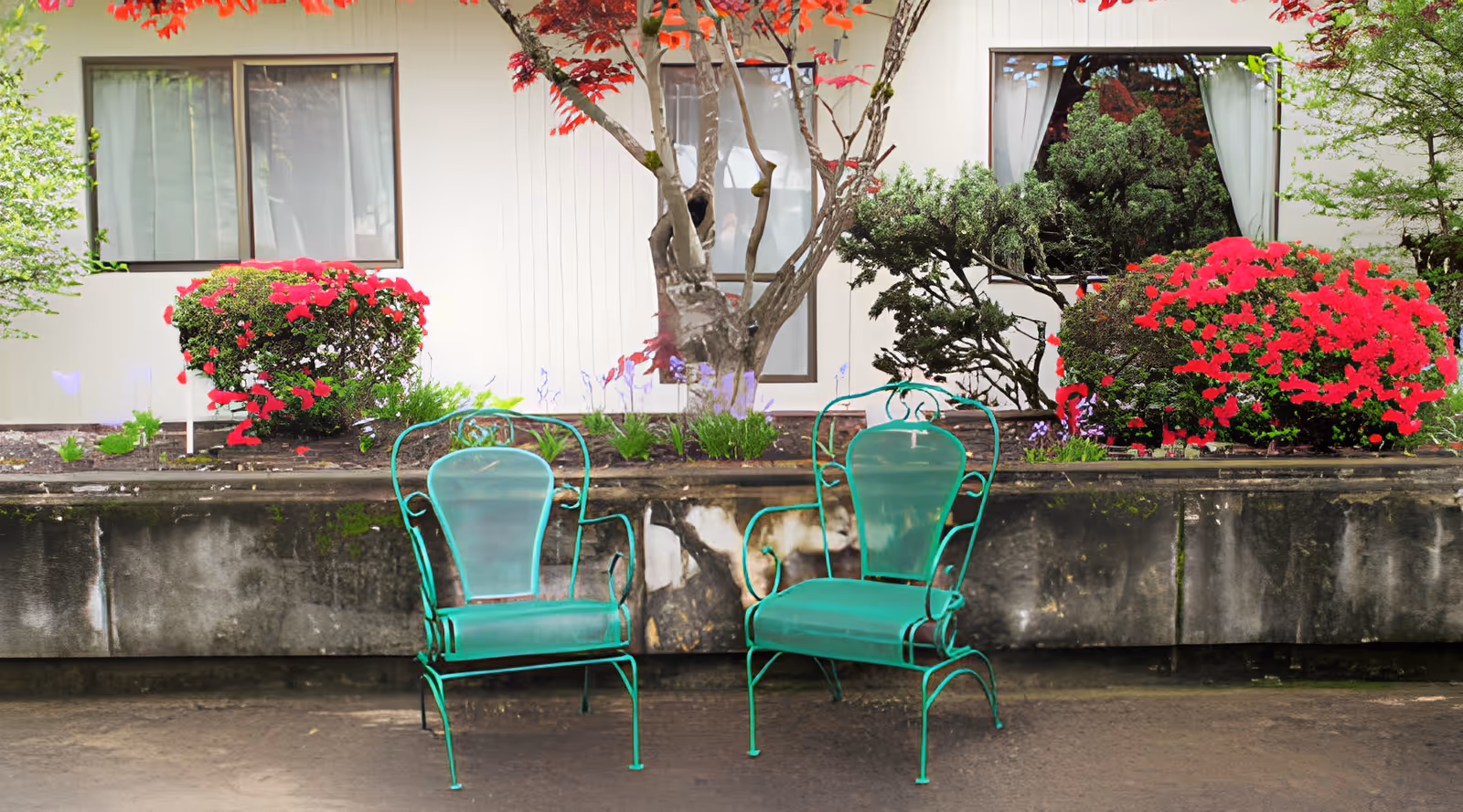 Two green metal chairs on a paved area in front of a low planter with flowering shrubs and a building facade with windows.