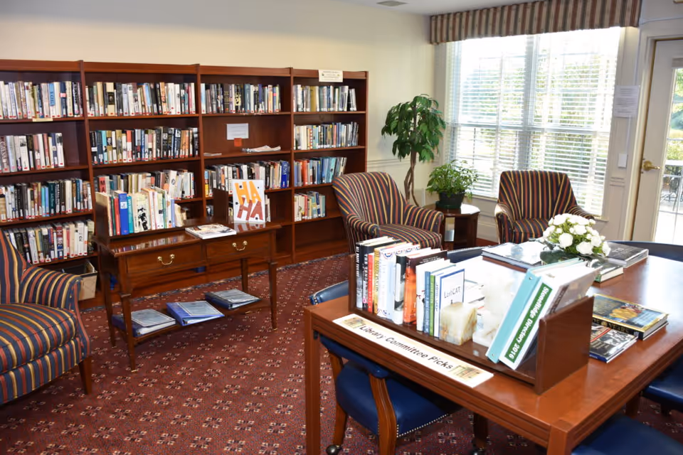 A cozy senior living facility library room with wooden bookshelves filled with books, a small wooden table with books and magazines, two striped armchairs, a large window with blinds letting in natural light, a potted plant, and a wooden table labeled 'Library Committee Picks' displaying a selection of books and a small flower arrangement.