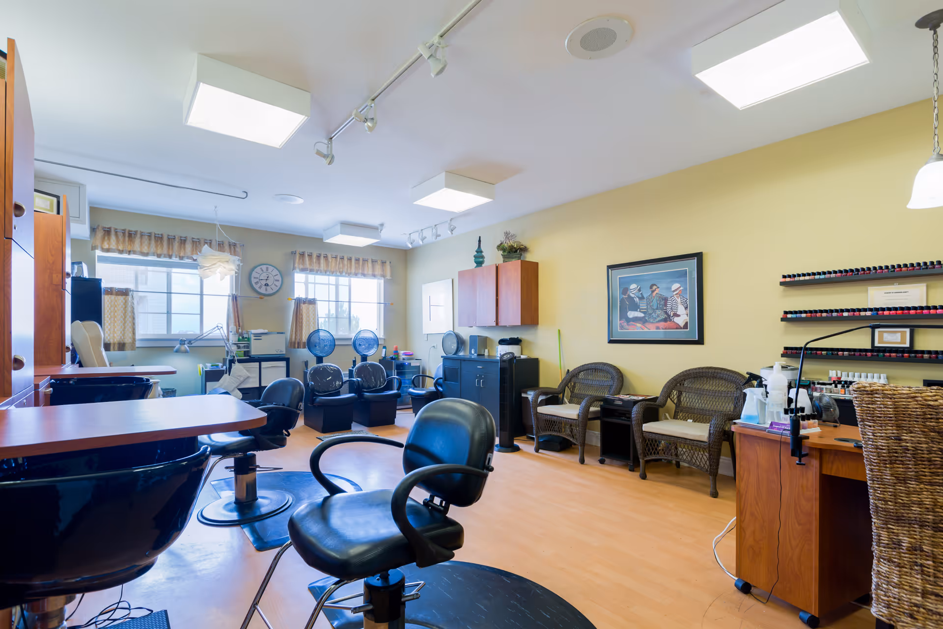 Interior view of a senior living facility's salon area with multiple black salon chairs, hair drying stations, wicker chairs along the wall, a nail polish display rack, and a framed picture on a yellow wall. The room has large windows with curtains and bright ceiling lights.