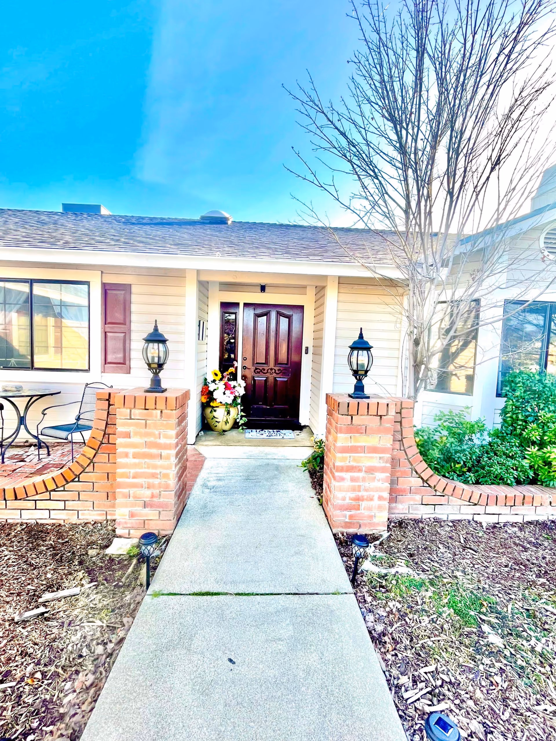 Front entrance of a single-story home with a concrete walkway flanked by low brick pillars, lanterns, a wooden front door and potted flowers.