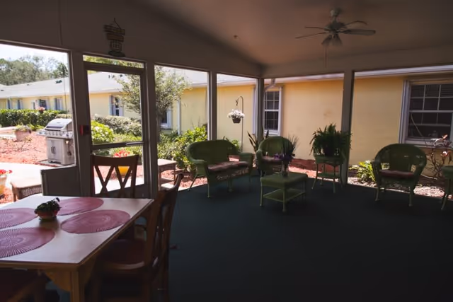 A covered patio area with green wicker chairs and a small table, a ceiling fan above, and a dining table with chairs and pink placemats in the foreground. Outside the screened patio, there is a garden area with plants and a barbecue grill, and a yellow building with windows in the background.
