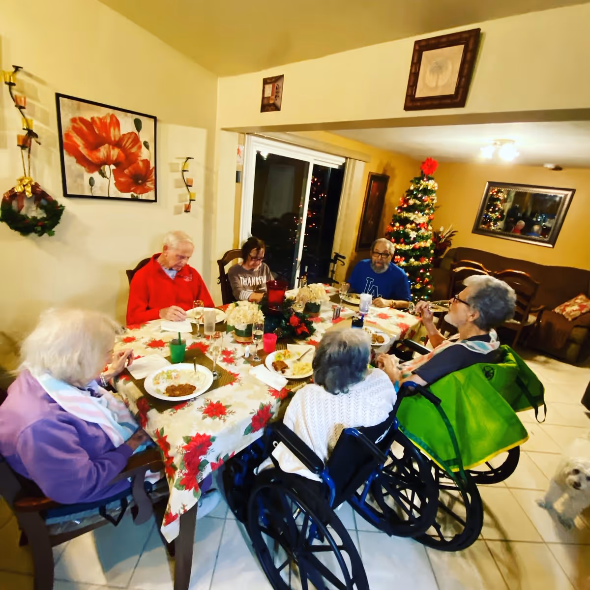 A group of elderly people sitting around a dining table decorated with a festive tablecloth and holiday centerpiece, enjoying a meal together in a warmly lit room with a decorated Christmas tree in the background. Two individuals are in wheelchairs, and a small white dog is visible on the floor nearby.