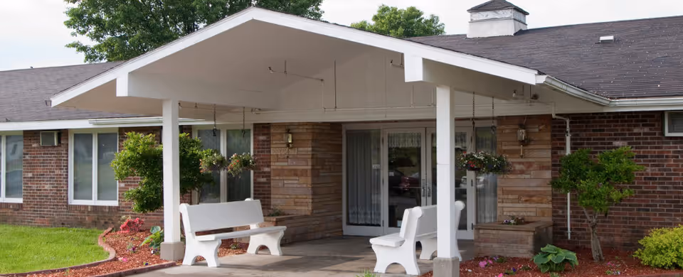 Entrance area of Prairie Village Nursing & Rehabilitation featuring a covered porch with white benches, hanging flower baskets, and a brick exterior building surrounded by green grass and small trees.