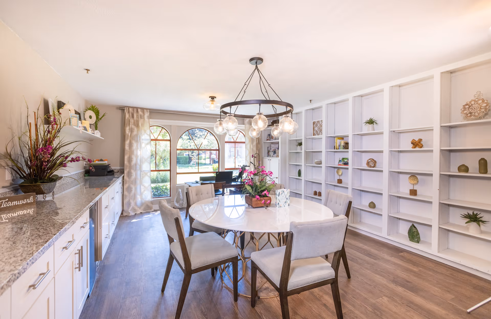 A bright and spacious interior room with a round white table surrounded by six cushioned chairs. Above the table hangs a modern chandelier with multiple globe lights. The room features a long countertop with cabinets underneath on the left side, decorated with plants and small items. On the right side, there is a large built-in white bookshelf with various decorative objects. Large arched windows at the far end let in natural light and offer a view of greenery outside.