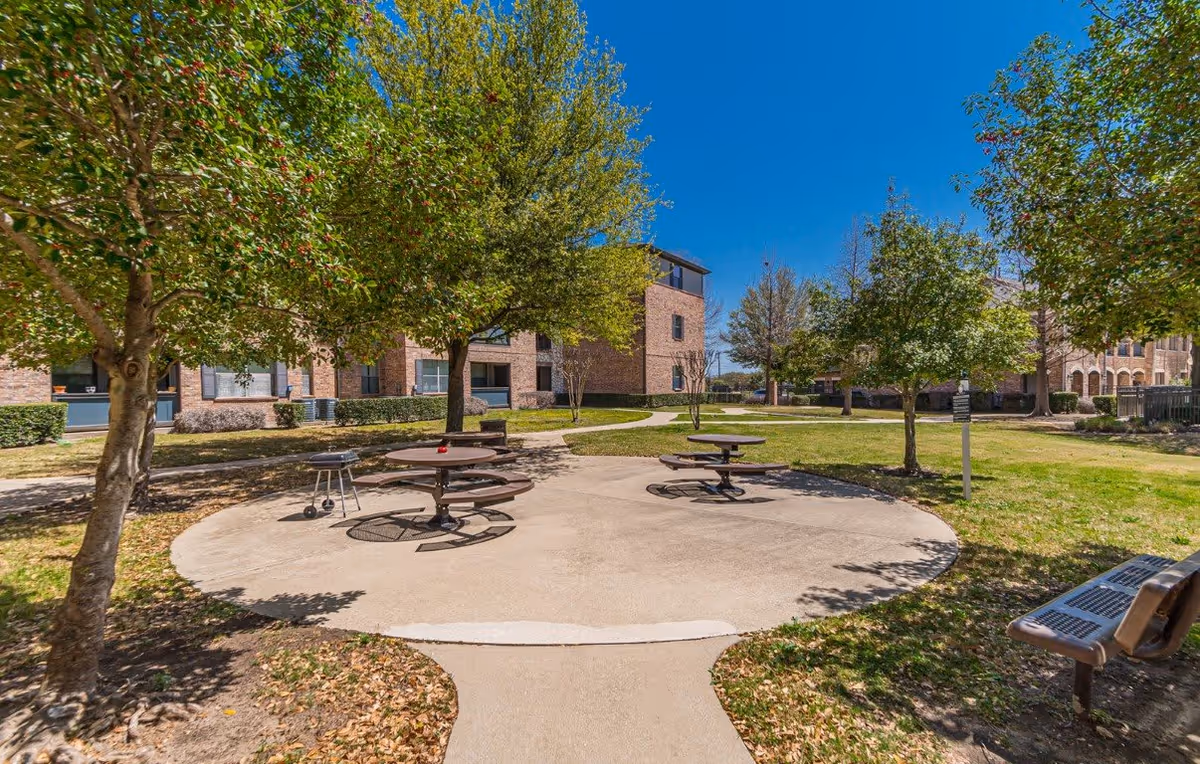 Outdoor seating area at The Cesera 55+ Apartments featuring round picnic tables with attached benches on a concrete circular patio, surrounded by green grass, trees, and a brick apartment building under a clear blue sky.