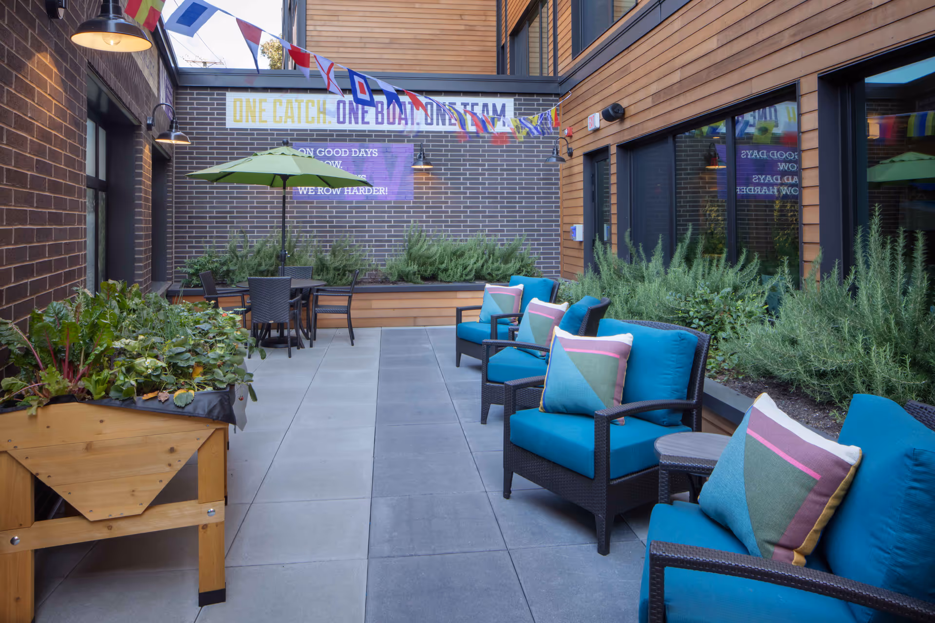 Outdoor patio area with blue cushioned chairs and colorful pillows arranged along the right side, a table with a green umbrella and chairs in the background, raised garden beds with plants on the left and right sides, and a brick wall with a motivational banner and colorful flags hanging above.