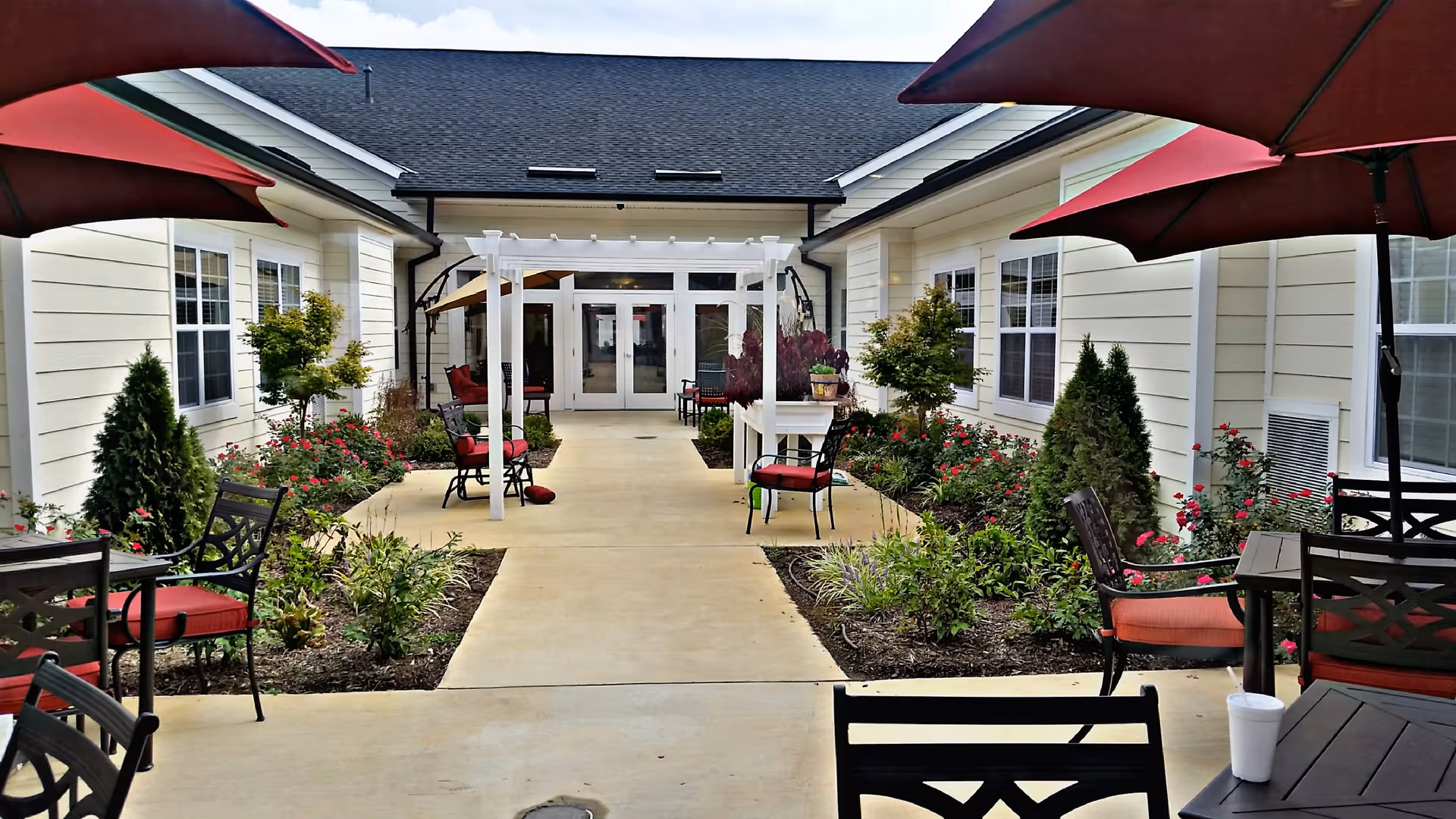 Outdoor courtyard area at Morningside House of Fredericksburg featuring a paved walkway lined with flower beds and shrubs. There are several black metal chairs with red cushions and tables under large red umbrellas. A white pergola structure is positioned along the walkway leading to glass double doors of the building.