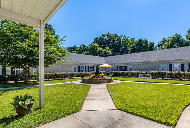 A sunny outdoor courtyard at Rock Hill Grove Senior Living featuring a circular flower bed with an umbrella in the center, surrounded by green grass, a paved walkway, a bench, and a single-story building with white siding and multiple windows in the background.