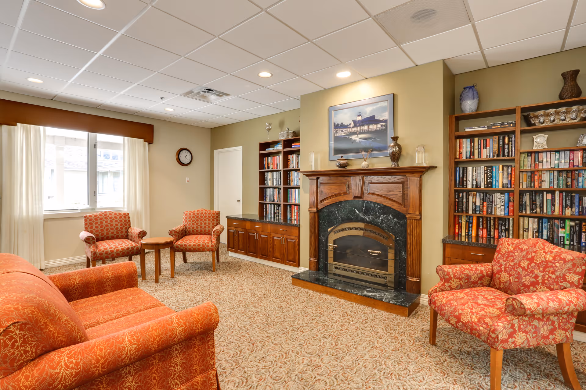 A cozy living room with patterned carpet, a wooden fireplace with a dark marble surround, and a framed picture above it. The room has built-in wooden bookshelves filled with books and decorative items. There are three upholstered chairs and a sofa in warm red and orange tones, along with a small wooden round table. A window with white curtains lets in natural light, and a wall clock is mounted on the beige wall.