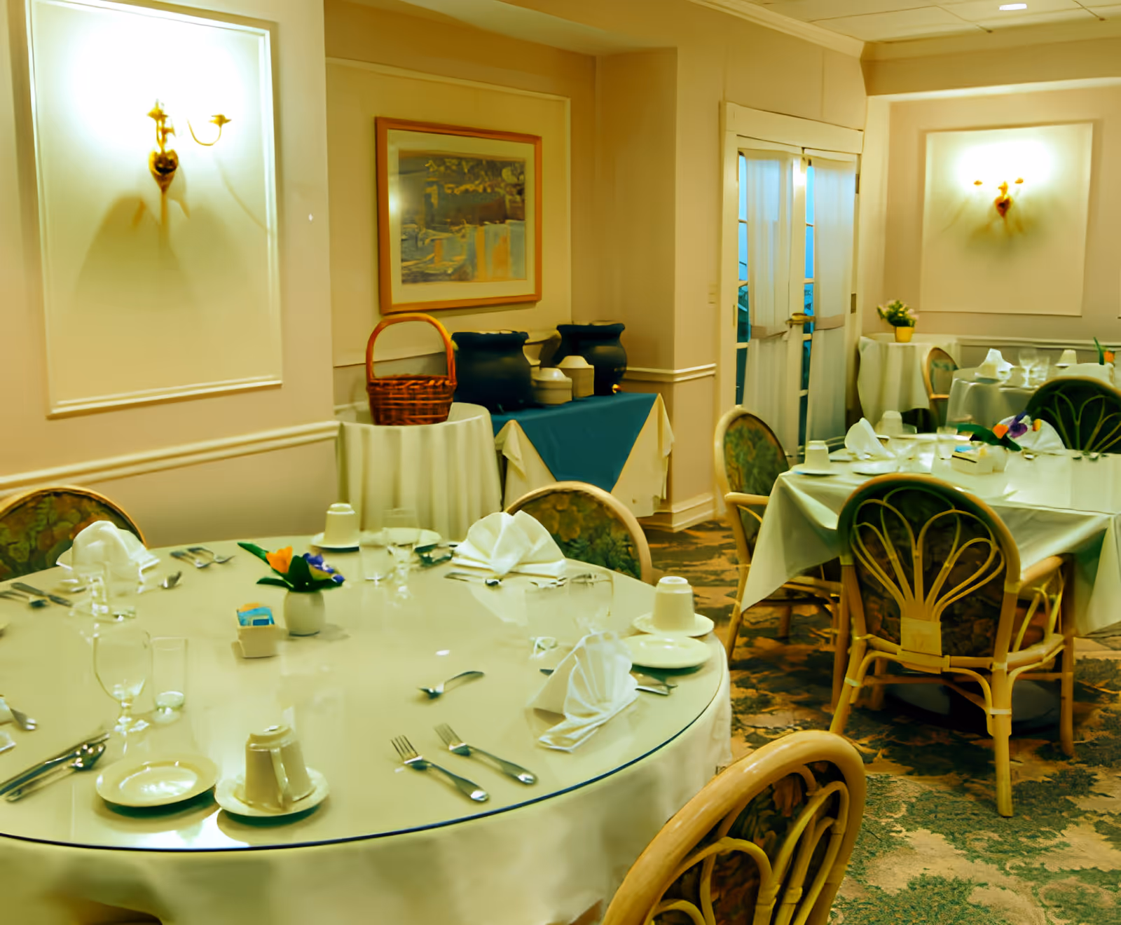 A dining room in Alden Town Manor Rehab & Health Care Center with round tables set with white tablecloths, folded napkins, glassware, and cutlery. The room has cushioned chairs with floral upholstery, wall sconces providing warm lighting, a framed painting on the wall, and a side table with beverage dispensers and a basket.
