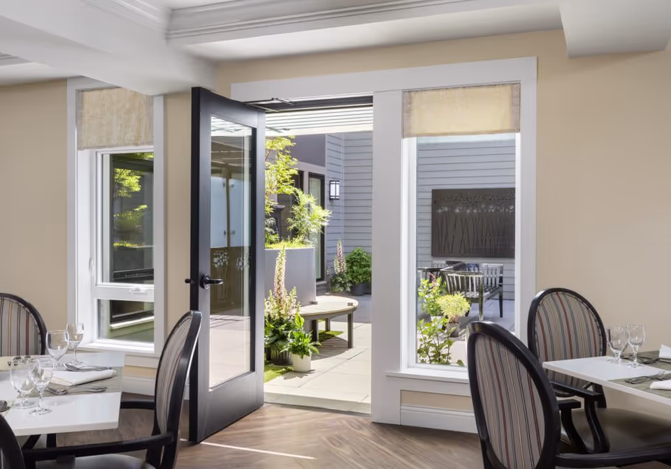 A bright dining area with tables set with glasses and napkins, featuring striped chairs. A black-framed glass door is open, leading to an outdoor patio with potted plants and seating. Large windows allow natural light to fill the space.