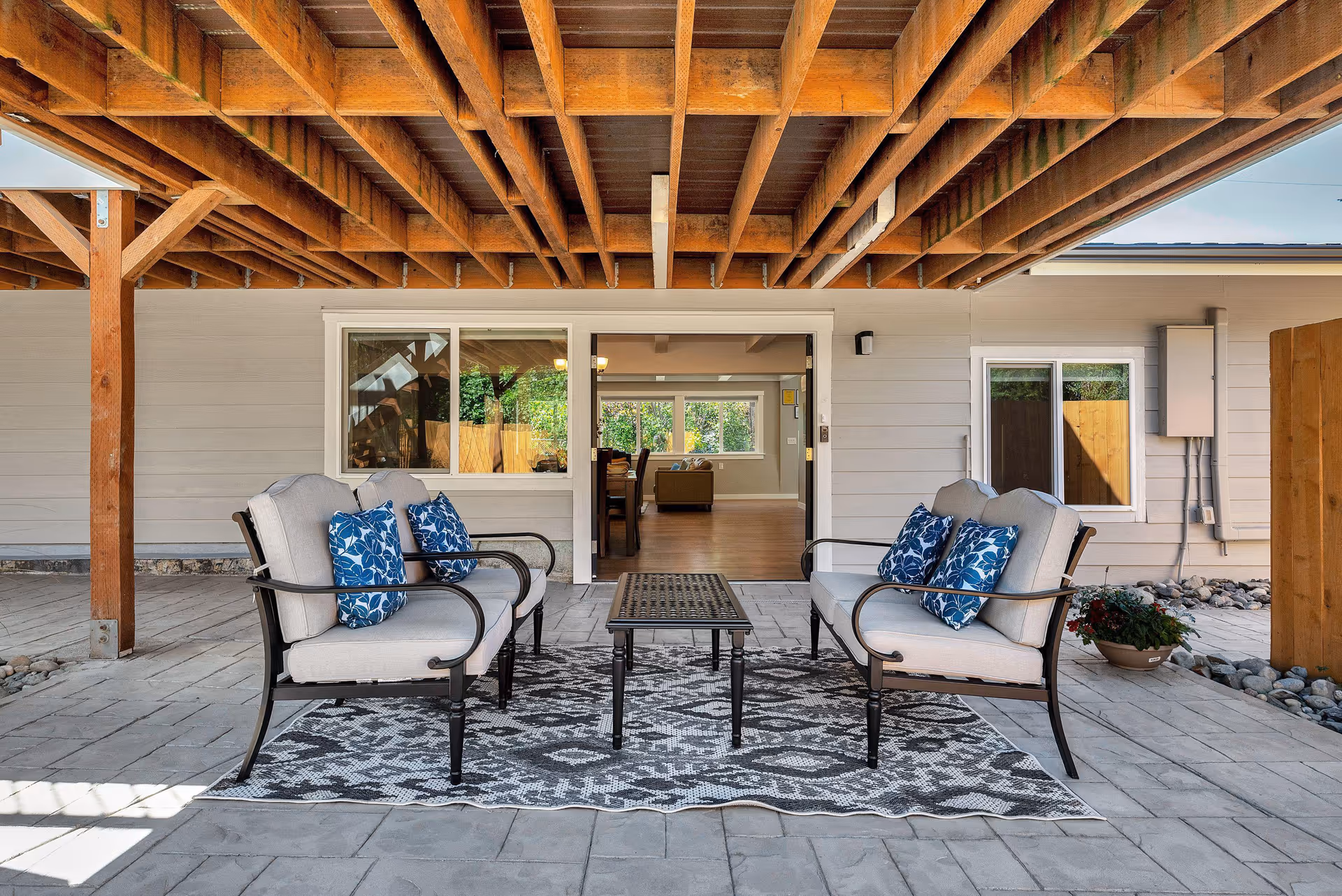 Covered patio under a wooden deck featuring two cushioned armchairs with blue pillows and a small table on a patterned rug in front of sliding doors into the house.