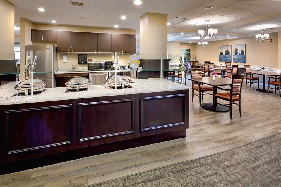 A spacious dining area in a senior living facility featuring a buffet counter with covered serving dishes and a glass sneeze guard. Behind the counter is a kitchen area with stainless steel appliances and wooden cabinets. The dining area has multiple tables and chairs, with framed artwork on the walls and chandeliers hanging from the ceiling.