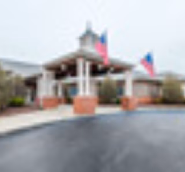 Exterior view of a single-story building with a covered entrance supported by white columns on brick bases. Two American flags are displayed near the entrance, and there is a paved driveway in front of the building.