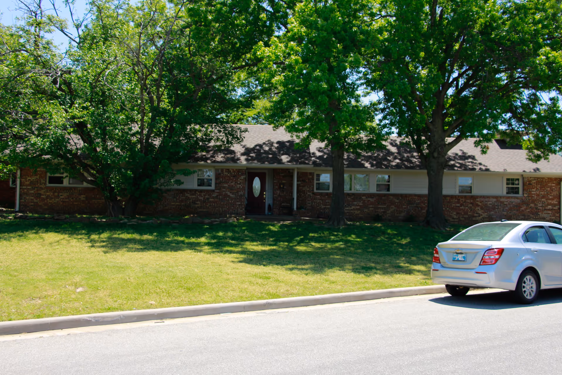 A single-story brick building with a dark shingled roof partially shaded by large green trees in front. A silver car is parked on the street in front of the building, which has several windows and a central front door with an oval glass panel.