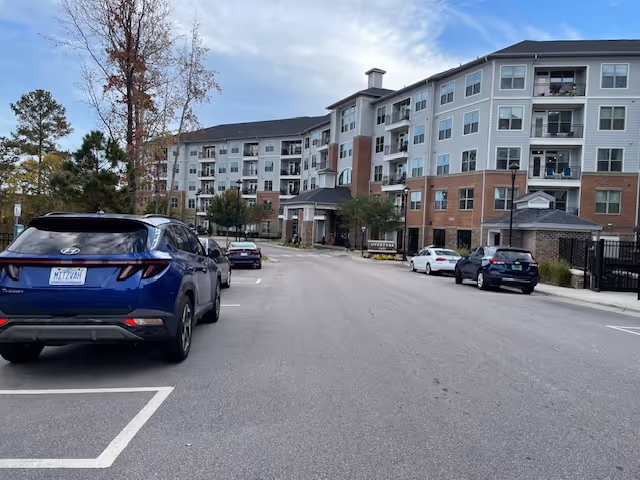 Overture Cary multi-story senior living building with a front driveway, parked cars, and trees under a partly cloudy sky.