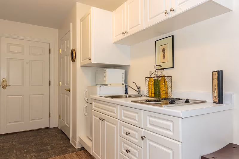 Small kitchen area with white cabinets, a sink, a microwave, a two-burner stovetop, and decorative bottles on the counter. There is a closed white door and a closet door in the background.