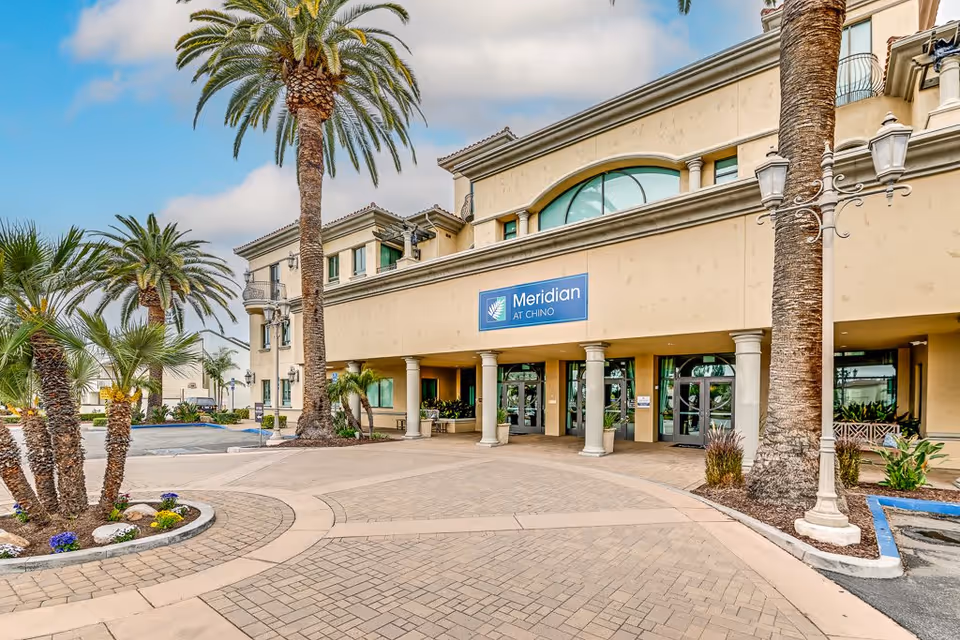 Exterior front view of Meridian at Chino senior living facility with palm trees, a paved driveway, and a beige building featuring large windows and a blue sign with the facility name.