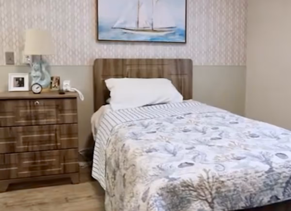 A neatly made single bed with a white pillow and a quilt featuring a seashell and coral pattern. Next to the bed is a wooden nightstand with a lamp, a clock, and framed photos. Above the bed hangs a painting of a sailboat on the water. The room has light-colored walls with a subtle patterned wallpaper behind the bed.