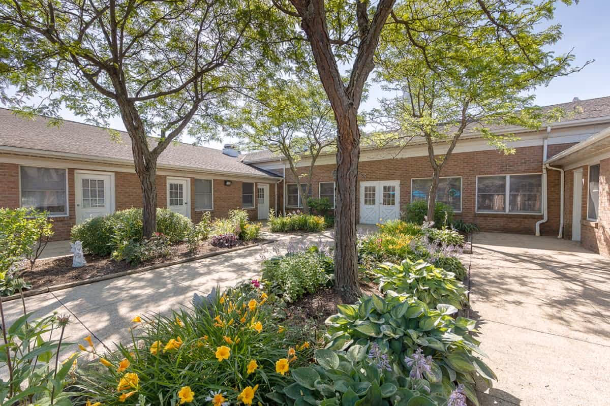 Sunlit courtyard with trees, flowerbeds and a paved walkway surrounded by single-story brick buildings with doors and windows.