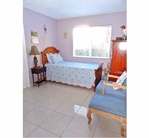 A lavender bedroom with a single bed, nightstand, lamp, window, and blue patterned chair.
