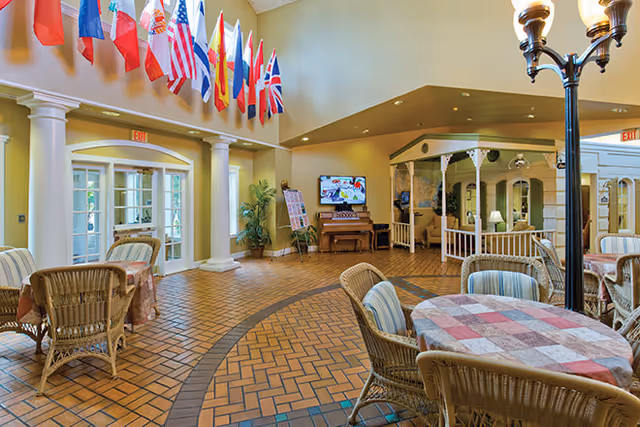 A spacious common area in a senior living facility with wicker chairs and tables covered with checkered tablecloths. The room features a tiled floor with a circular pattern, a piano against the far wall, and a television mounted above it. Multiple international flags hang from the ceiling near large windows and glass doors. There is a cozy seating area with a white railing and decorative trim, and a tall lamp with multiple lights stands near the tables.