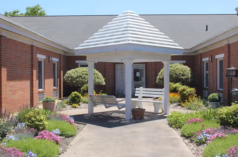 Outdoor courtyard area at Mayfair House Senior Living featuring a white gazebo with a swing and bench underneath, surrounded by colorful flower beds and shrubs, with brick building walls on either side.