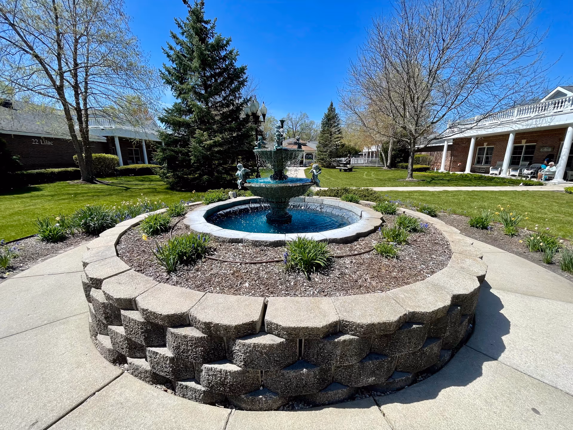 Outdoor courtyard area with a circular stone planter and a tiered water fountain in the center. Surrounding the planter are green lawns, trees, and brick buildings with white columns and porches. The sky is clear and blue.