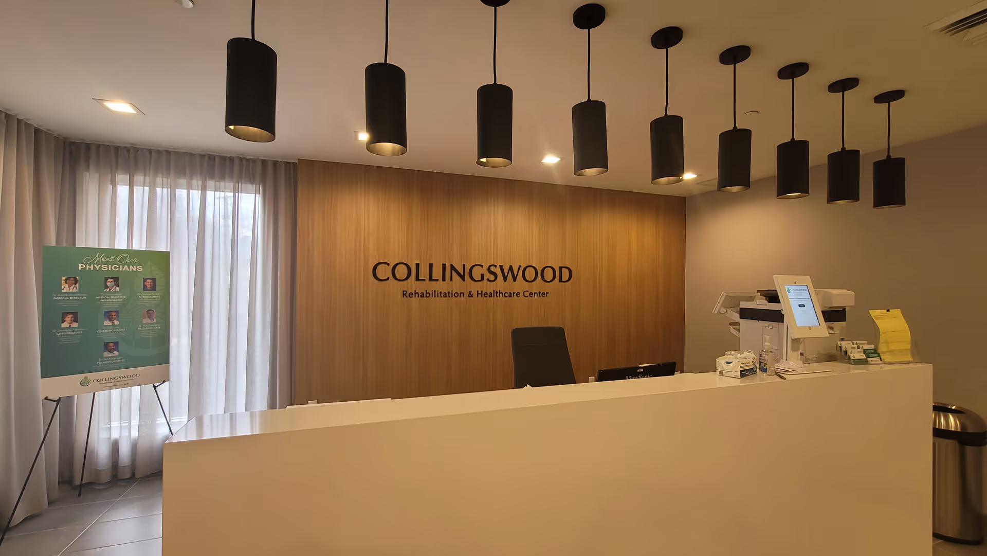 Reception area of Collingswood Rehabilitation & Healthcare Center with a white front desk, a black office chair behind it, and a wooden wall with the facility name. There are nine black pendant lights hanging from the ceiling. To the left, there is a green informational board about physicians next to a window with sheer white curtains.