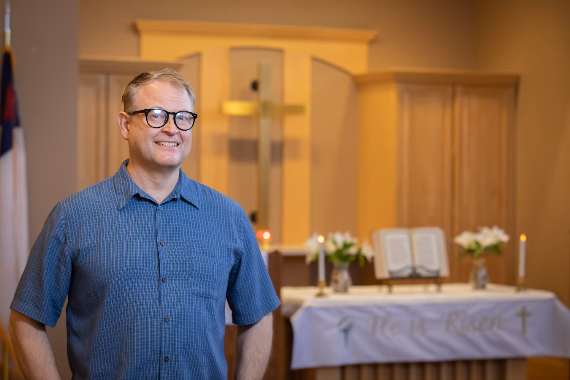 A middle-aged man wearing glasses and a blue checkered short-sleeve shirt stands smiling in a chapel or worship room. Behind him is an altar with a white cloth that has the words 'He is Risen' embroidered on it, along with a cross, candles, flowers, and an open book. A wooden cross is mounted on the wall in the background.