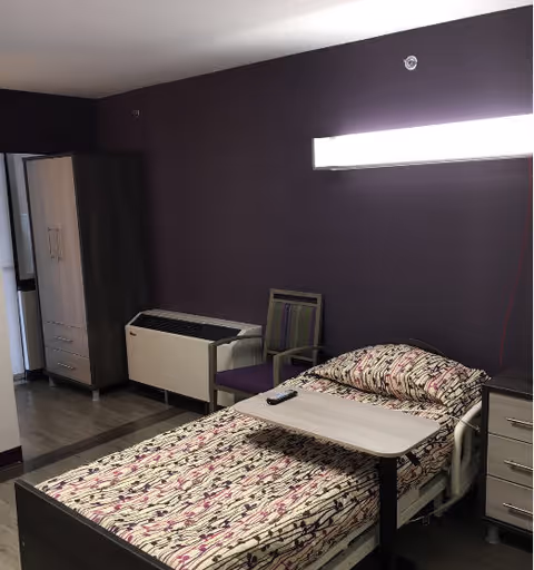 A resident bedroom with a hospital-style bed, patterned bedding, an overbed table, a chair, wardrobe and a purple accent wall.