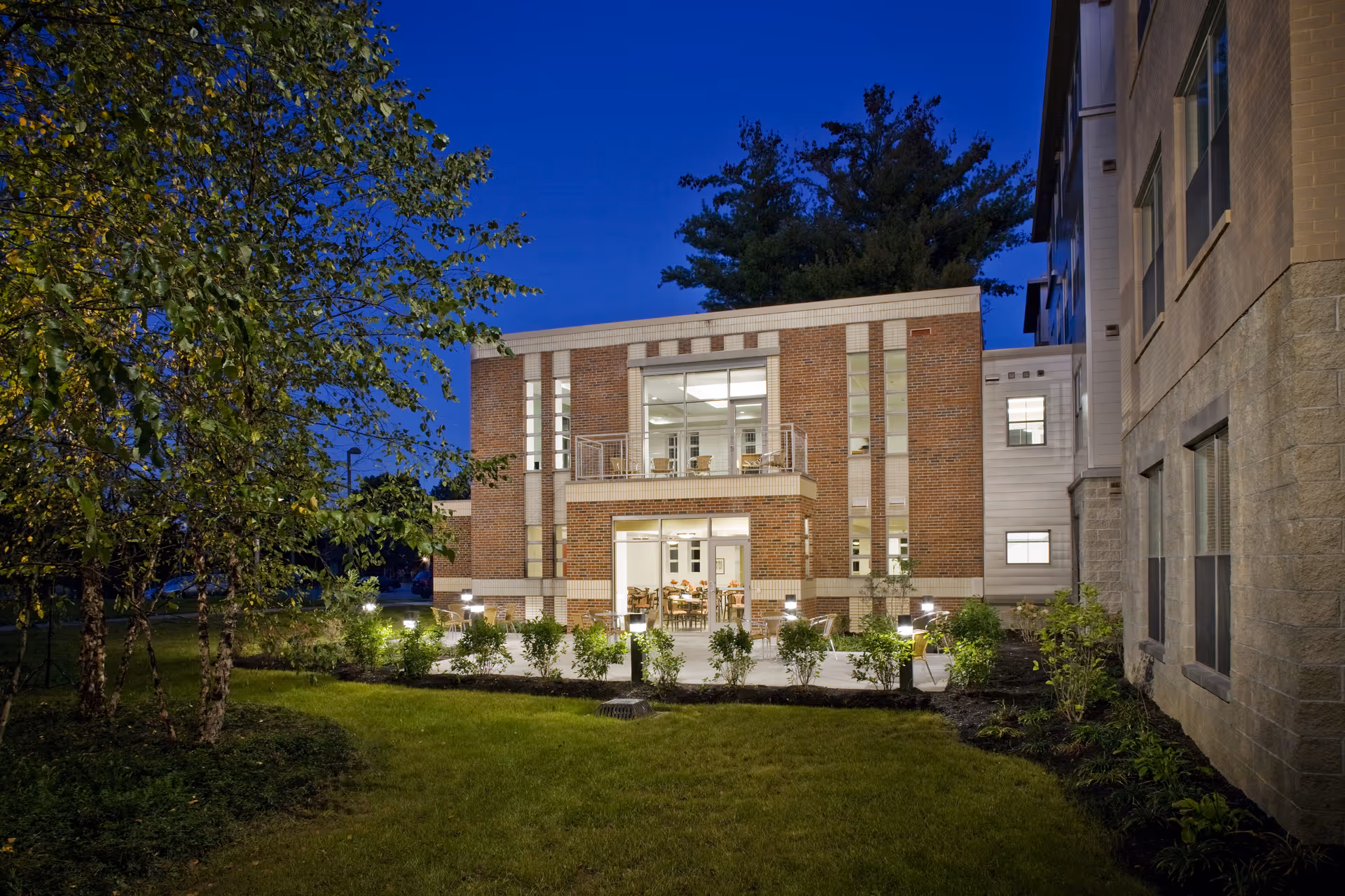 Exterior view of a modern brick and stone building at dusk with large windows showing a well-lit interior dining area. The building is surrounded by a green lawn, small bushes, and trees, with outdoor lighting illuminating the pathway.
