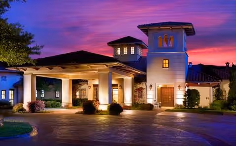 Exterior view of a large, well-lit building at dusk with a purple and pink sky in the background. The building has a covered entrance with columns, multiple windows, and a tower-like structure on the right side. There are shrubs and trees around the building and a paved driveway in front.