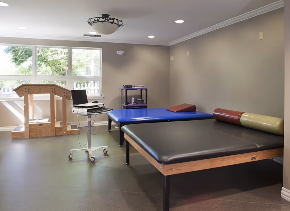 A therapy or rehabilitation room with two padded therapy tables, one black and one blue, each with cylindrical and wedge-shaped bolsters. There is a wooden stair-step apparatus near a large window letting in natural light. A mobile computer workstation with a laptop is positioned near the tables, and a small shelving unit with supplies is against the wall.