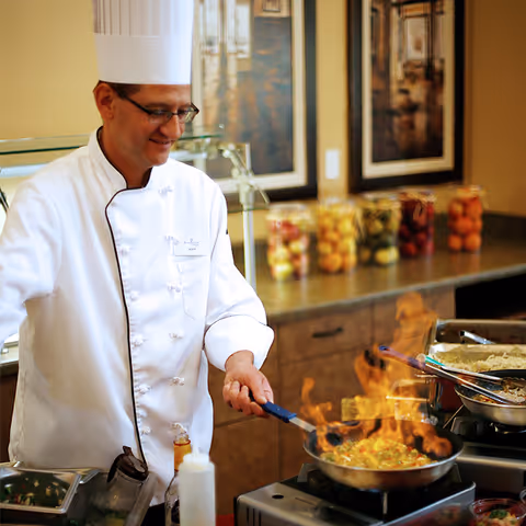 A chef wearing a white uniform and tall white hat is cooking with a pan over a stove, with visible flames. The kitchen counter behind him has jars filled with various colorful ingredients and framed pictures on the wall.