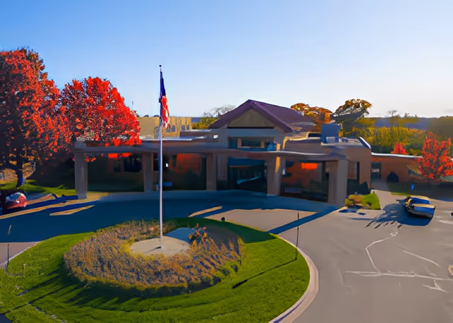 Front exterior view of Martin Luther Campus building with a circular driveway, a flagpole with an American flag in the center of a landscaped roundabout, and trees with red autumn foliage in the background under a clear blue sky.