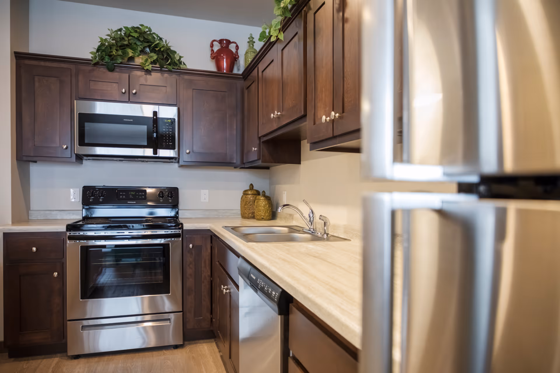 Modern kitchen with stainless steel appliances including a refrigerator, oven, microwave, and dishwasher. The kitchen features dark wooden cabinets, a light-colored countertop, a double sink, and decorative plants and jars on top of the cabinets.