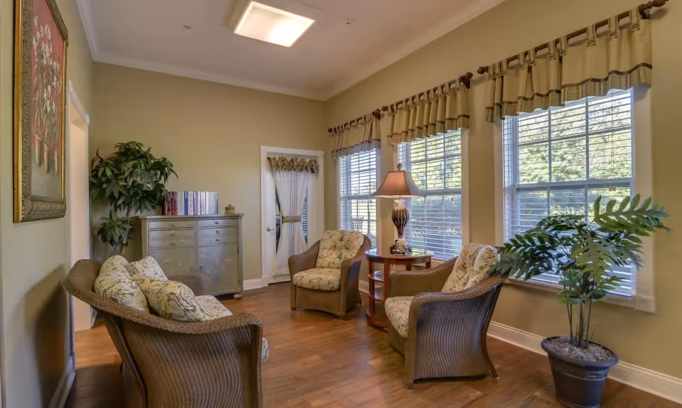 Sunlit sitting room with four wicker armchairs around a lamp-topped side table, hardwood floors, large windows, and potted plants.