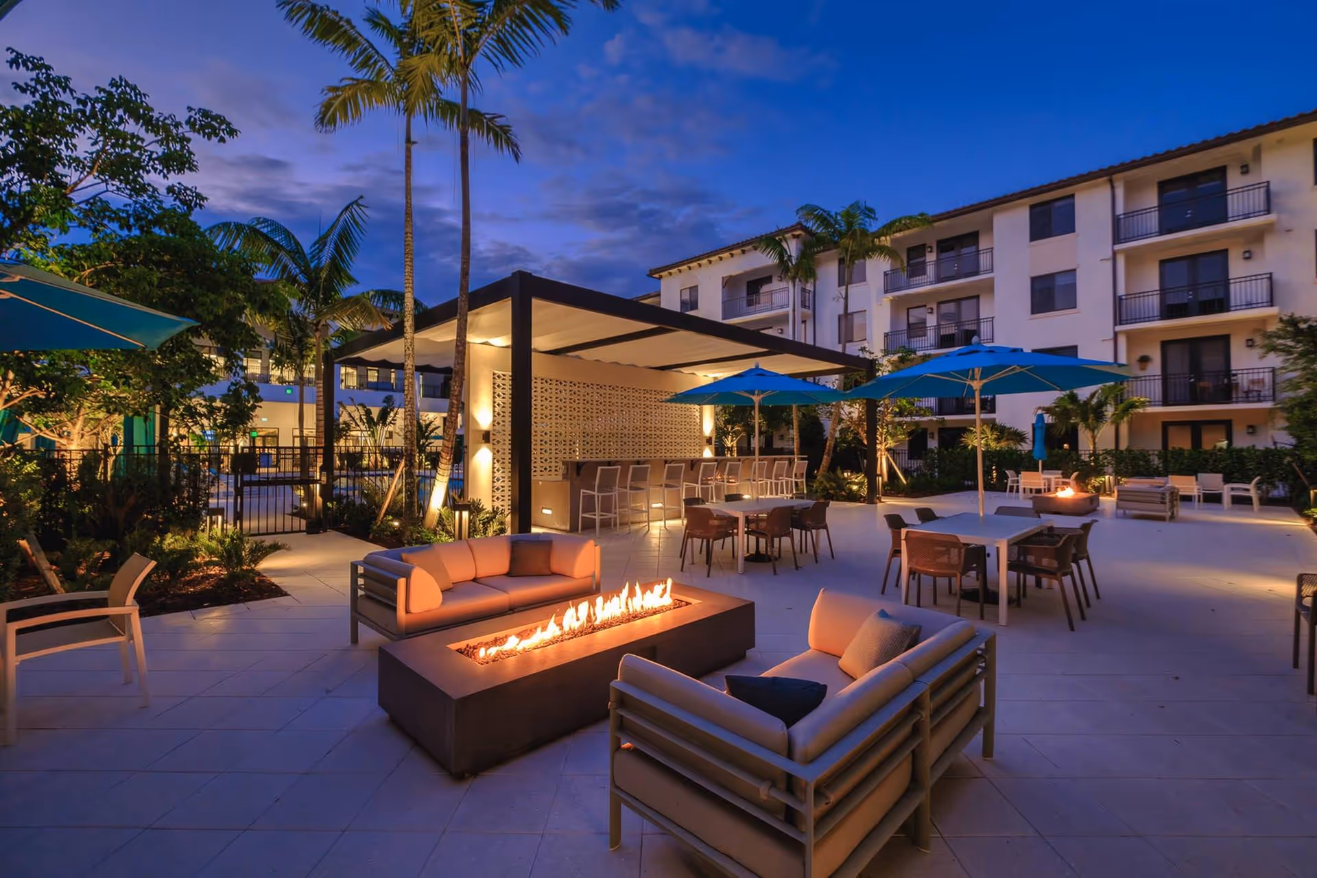 Dusk view of an outdoor courtyard with lounge seating, a long fire pit, dining tables with umbrellas, and a multi-story residential building in the background.