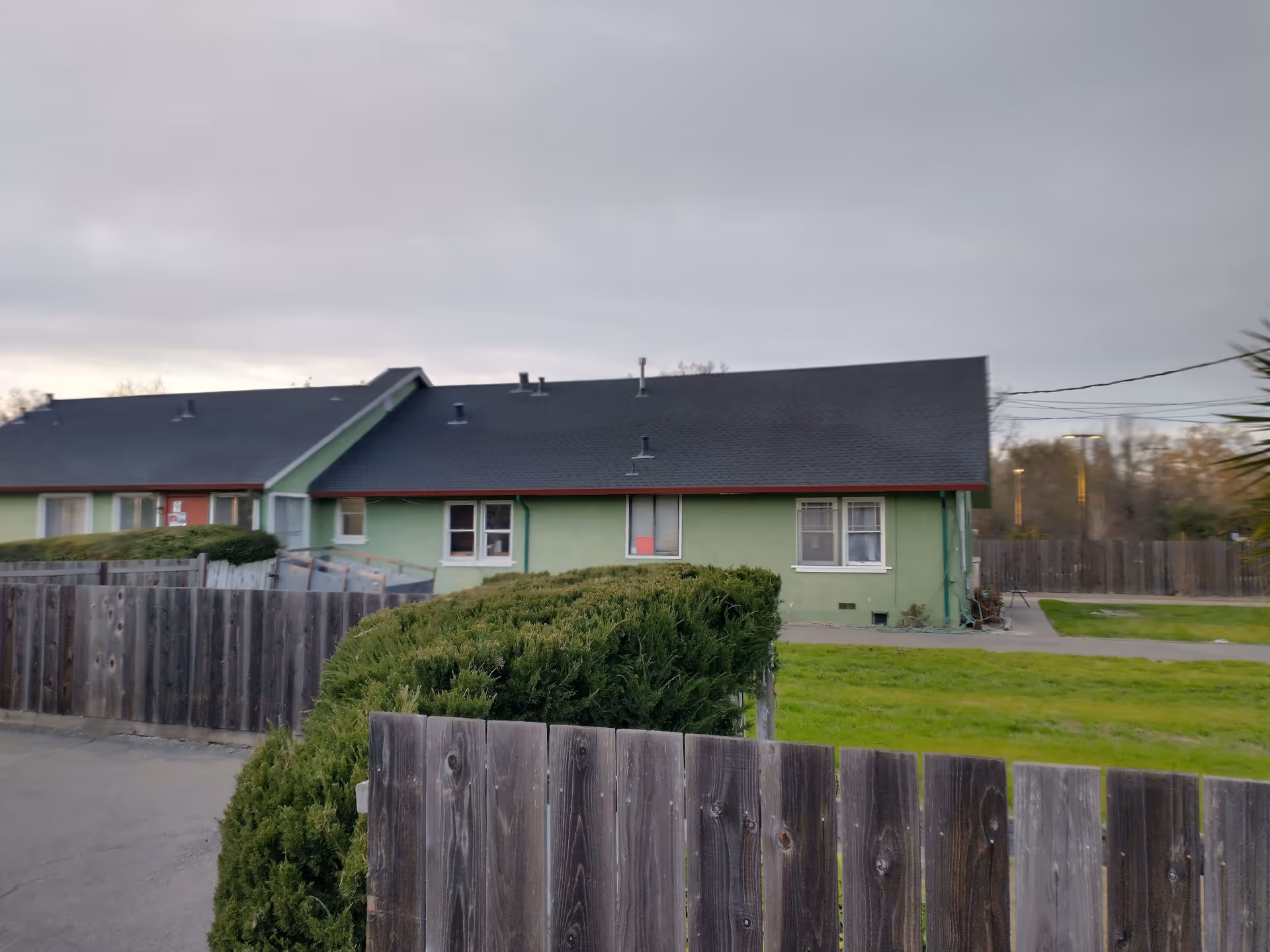 Exterior view of a single-story green building with a dark roof, surrounded by a wooden fence and green bushes, under a cloudy sky.