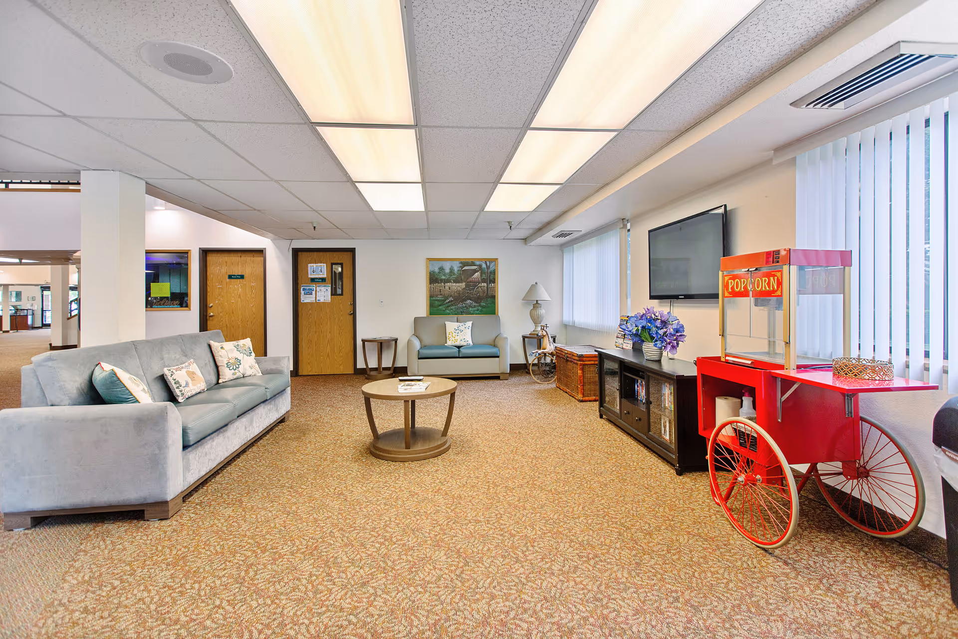 A spacious common living area with a gray sofa and loveseat, a round wooden coffee table, a black entertainment cabinet with a flat-screen TV mounted above it, and a red popcorn cart. The room has beige patterned carpet, white walls, and large windows with vertical blinds letting in natural light. There are two wooden doors on the far wall and a framed painting above the loveseat.