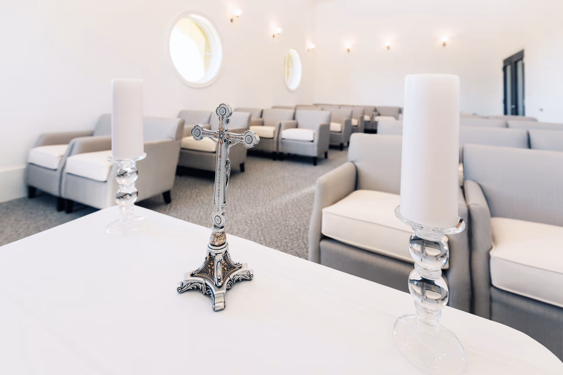 Silver cross and two white candles on a table in front of rows of gray upholstered chairs in a bright chapel-like room.