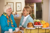 Two elderly women sitting at a table in a cozy indoor setting, smiling and enjoying conversation. The table has a tray with croissants, apples, and oranges, and there are cups of coffee or tea in front of them. The background shows a comfortable living area with framed pictures on the wall.