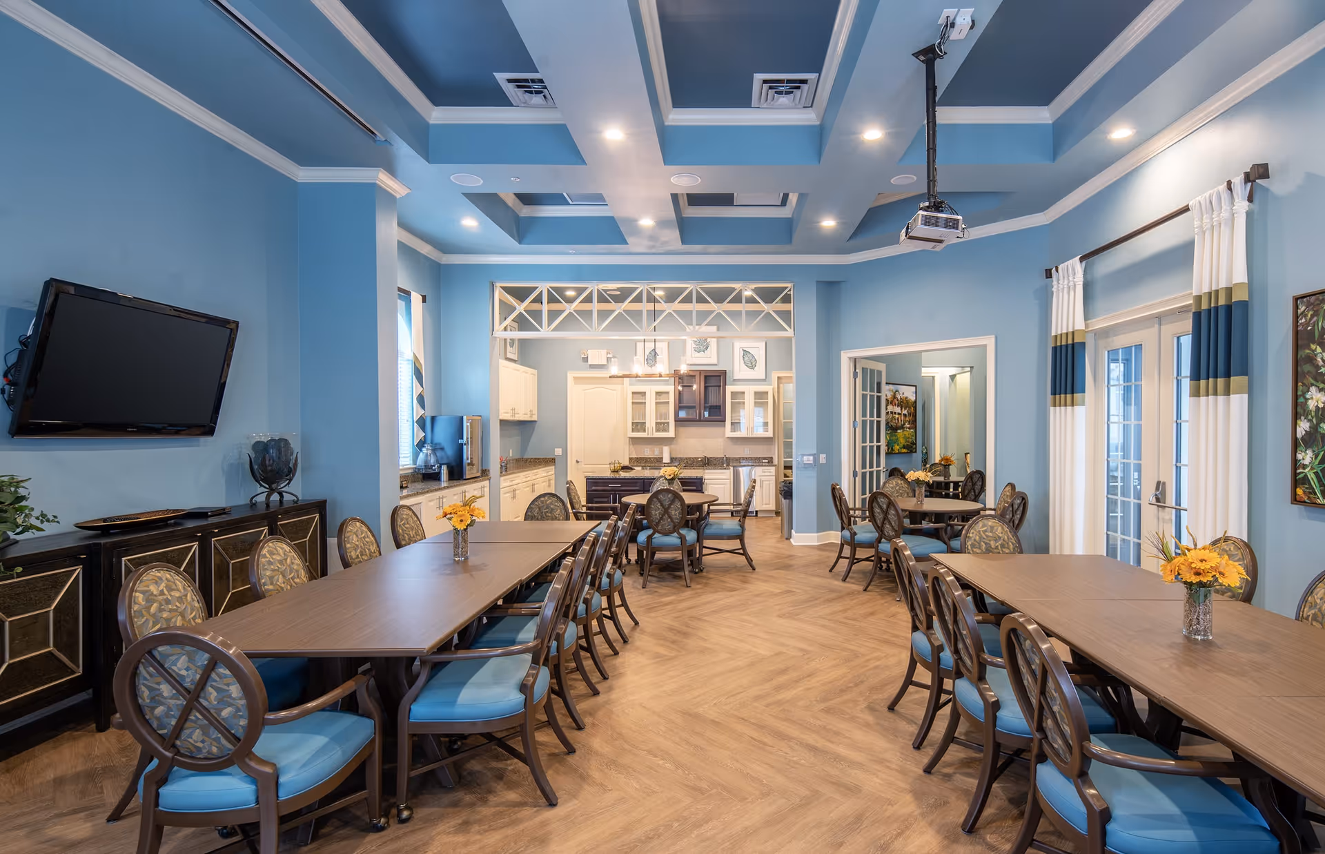 A spacious dining room with blue walls and ceiling, featuring two long wooden tables surrounded by chairs with blue cushions. The room has a mounted flat-screen TV, decorative curtains on the windows, and a kitchen area visible in the background. The floor has a wood pattern, and there are small flower arrangements on the tables.