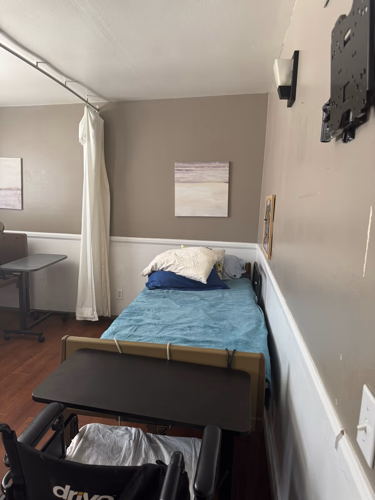 A small senior-care bedroom with a single adjustable bed made with blue bedding, an overbed table, a wheelchair, wall art, and a privacy curtain.